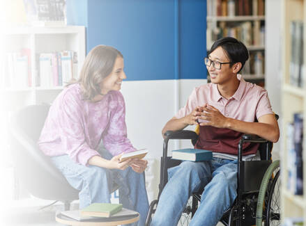 Two teenagers in a library talking with each other. One teenager is in a wheelchair.